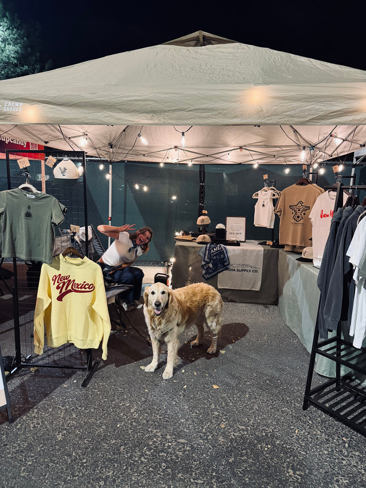 The Sandia Supply Co. market booth at night under string lights, with tees and a New Mexico sweatshirt on racks and a golden retriever out front